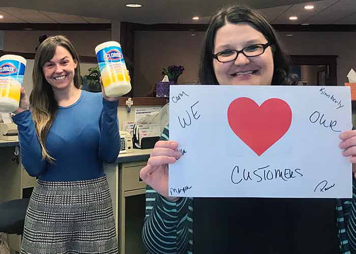 Two bankers: one holding up disinfectant wipes, one holding a sign that says, "We (Heart) Our Customers"